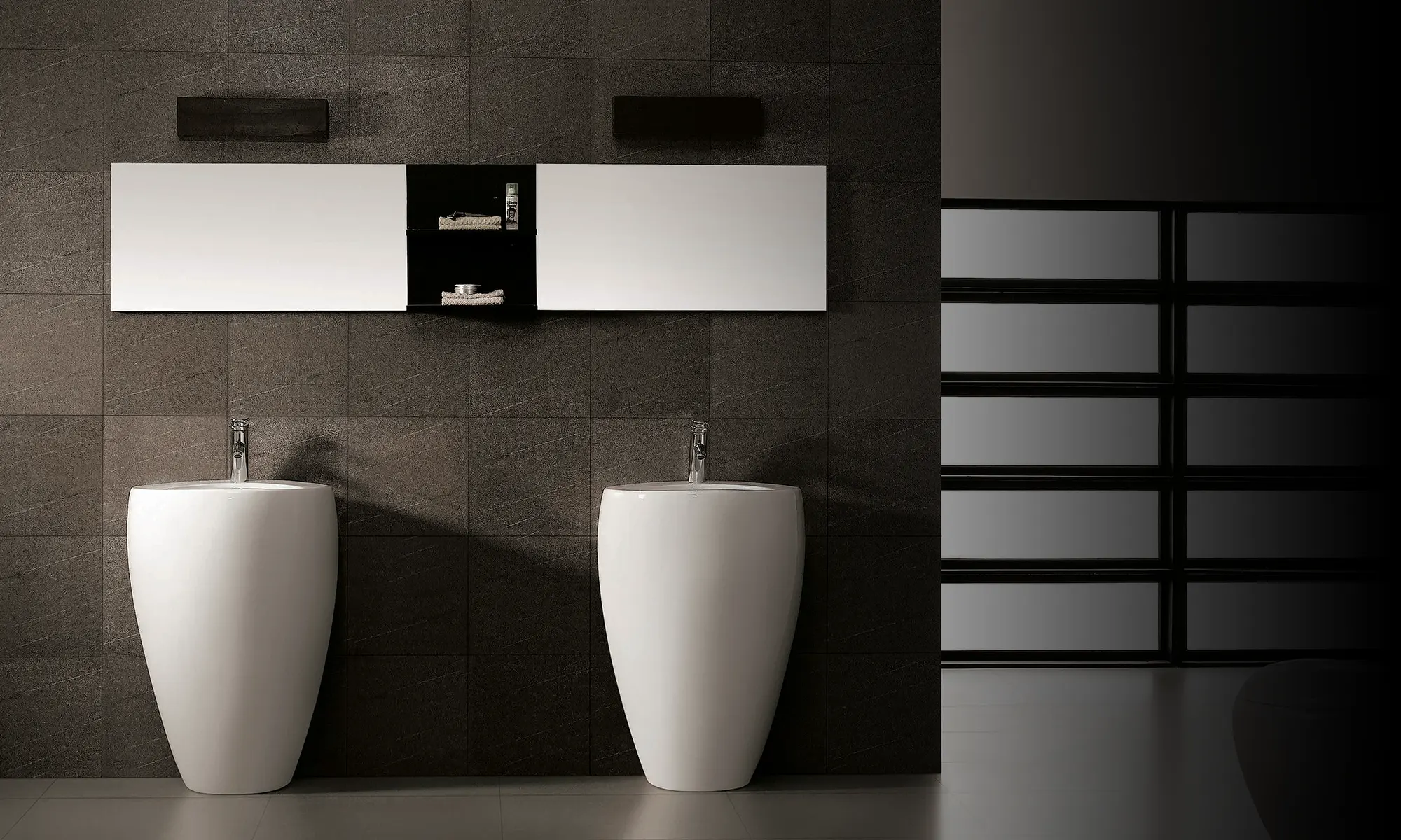 Two white freestanding pedestal wash basins with chrome pillar taps in a dark tiled bathroom